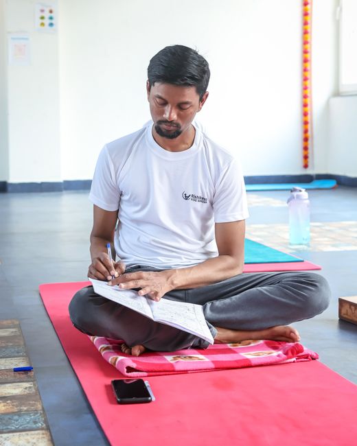 A man sitting cross legged on a mat and writing in a journal during a morning ritual.